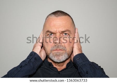 Determined man covering his ears with his hands to block out something he does not wish to hear or in a concept of hear no evil staring at the camera on a grey background