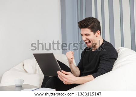 Image of a happy handsome young man at home indoors using laptop computer make winner gesture.