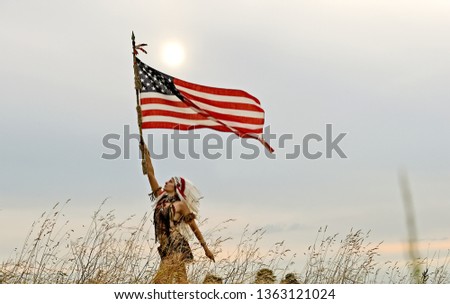 A young woman dresses up as an Indian warrior. 
She wears a white feathered bonnet on her head.
She stands outdoors in nature proudly holding a
US flag in her hand. 