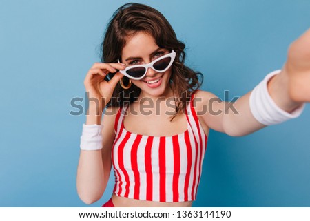 Attractive brunette girl takes off her glasses and coquettishly looks into camera. Lady in red and white top makes selfie on blue background