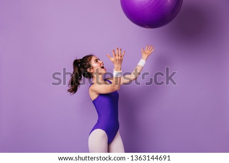 Pretty girl athlete in purple sports bodysuit in style of 80s throws fitball on isolated background