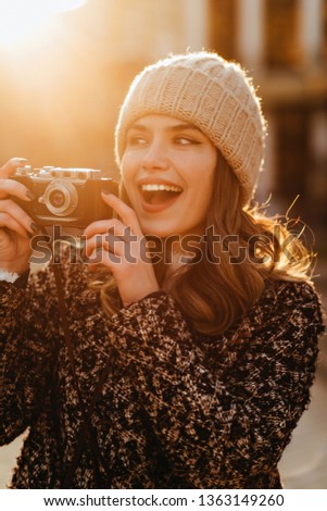 Well-dressed laughing woman with wavy hair holding camera. Charming female photographer working in sunny autumn day.