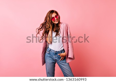 Excited white girl in bright stylish glasses posing on pink background. Dreamy curly woman playing with her ginger hair and laughing.