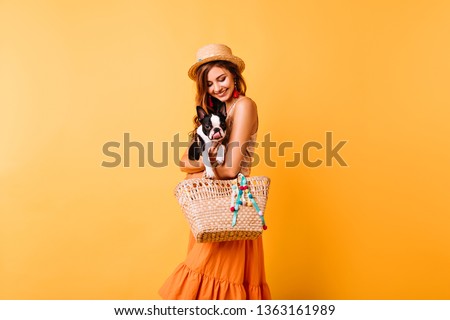 Pretty white girl in elegant hat embracing black bulldog. Indoor photo of smiling female model in bright attire posing with puppy.