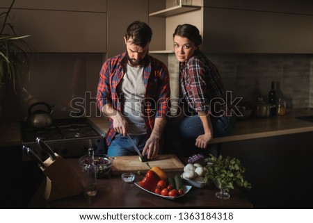 Beautiful young couple in kitchen at home while cooking healthy food. Man is cuts salad. Woman sits on table and looks in frame. Scene from family life. Horizontally framed shot.