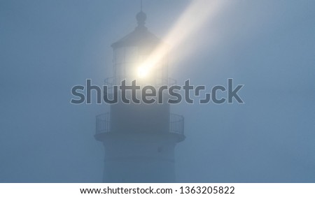 A beam of light from the Portland Headlight can be seen shining through heavy midst of fog in early morning hours off the coast of Maine. Rays of light from light house reflecting through fog in dark.