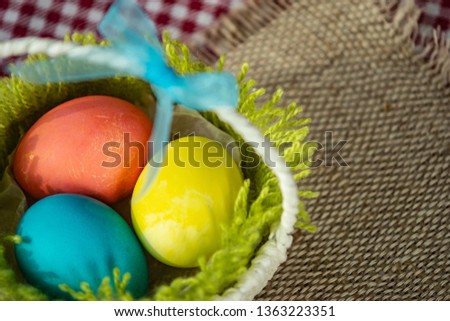 Top view closeup on colorful Easter eggs lying on green grass made of wool in a white basket with a blue ribbon, background of burlap and checkered tablecloth, place for text.