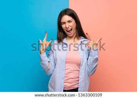 Young woman over pink and blue wall making rock gesture