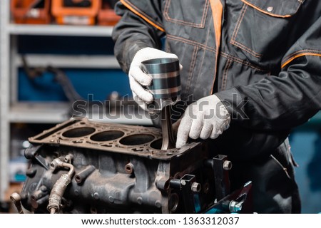 Car mechanic holding a new piston for the engine, overhaul.. Engine on a repair stand with piston and connecting rod of automotive technology. Interior of a car repair shop. Royalty-Free Stock Photo #1363312037