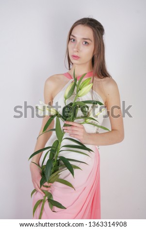 a charming young girl with big eyes and brown hair stands without makeup in a fashion dress on a white background in the Studio. beautiful young teen woman holding a stem with white lush lilies.