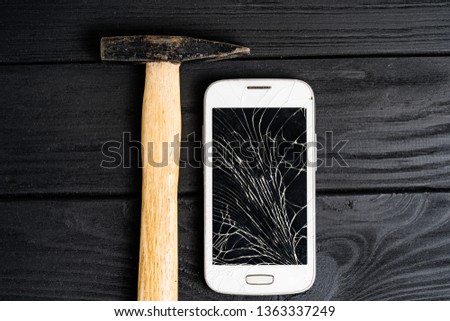Smartphone smashed by hammer isolated on black wooden background. Broken touchscreen of a smartphone lying close to the hammer on table.