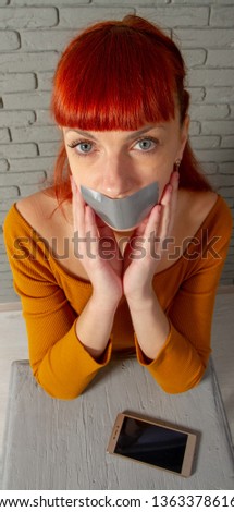 Conceptual picture. Young red-haired girl sitting near a smartphone with adhesive tape lips to keep silence