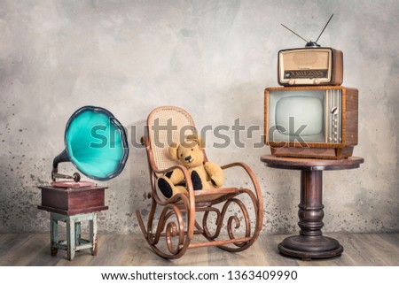 Retro outdated television, broadcast radio from circa 50s on wooden table,  old phonograph, Teddy Bear toy sitting on aged rocking chair front concrete wall background. Vintage style filtered photo 