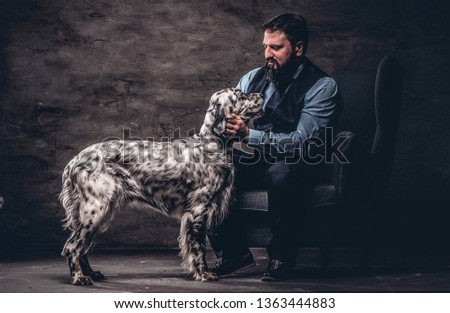 Bearded hunter wearing elegant clothes sitting on a sofa with his white English setter. Studio photo against a dark textured wall