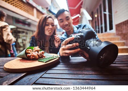Happy young chinese couple sitting at cafe outside enjoying traditional asian food and watching pictures in photo camera. Widescreen shooting.