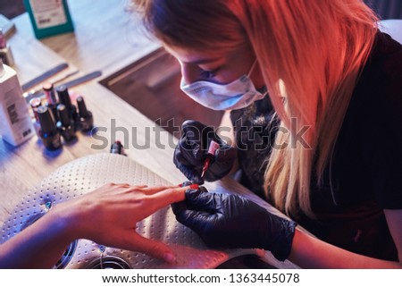 Top view of a beautician master applying polish on client's natural nails at the manicure table in the beauty salon. Photo with dim light and red Blue backlight