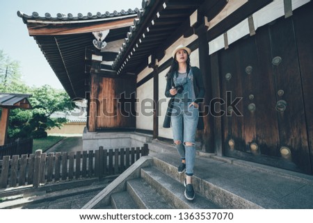 full length asian tourist woman is going down from stairs with camera outside of shitennoji temple osaka japan. young girl traveler walking with hands in pocket taking picture visit shinto looking.