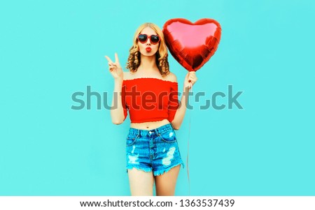 Pretty cool girl sending sweet air kiss with red heart shaped balloon on colorful blue background