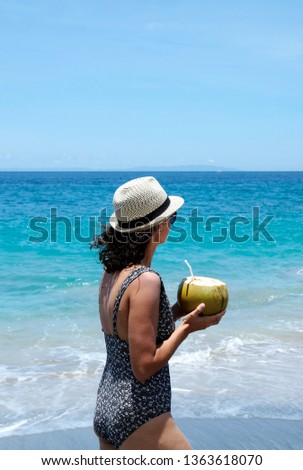 A woman wearing a hat and a swimming suit walking along a beautiful coastline and holding a huge drinking coconut. White sand beach, Bali, Indonesia