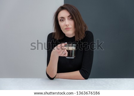 Conceptual portrait of a stylish brunette girl on a white and black background is drinking coffee while sitting at the table. Fashionable photo of beautiful girl with dark hair posing in the studio.