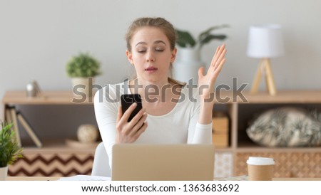 Irritated young woman using phone at workplace, reading bad news, receiving unpleasant email, conversation, feeling annoyed, looking at smartphone screen, sitting at home or office, making video call Royalty-Free Stock Photo #1363683692