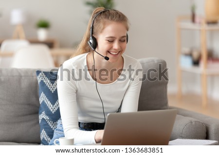 Smiling woman wearing headset with microphone, using laptop at home, sitting on sofa in living room, looking at screen, making video call, working support operator, translator, watching webinar