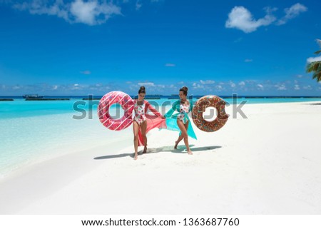Summer Vacation. Happy free two women with donut float mattress. Girls wearing Chiffon Beach Dress enjoying exotic beach by turquoise water seaside. Maldives island paradise background. 
