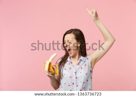 Crazy screaming young girl with eyes closed showing horns up gesture hold fresh ripe banana fruit isolated on pink pastel background. People vivid lifestyle relax vacation concept. Mock up copy space