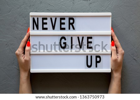 Female hands hold modern board with 'Never give up' words over gray background. Flat lay, from above, overhead. Close-up.