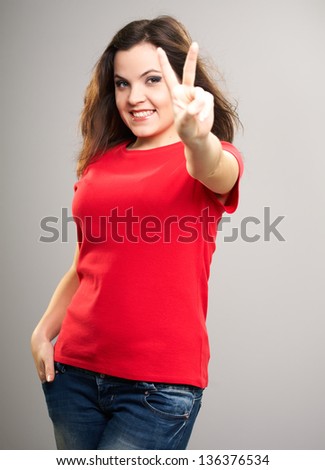 Attractive young woman in a red shirt. Woman shows a symbol of victory. Hair in motion. On a gray background