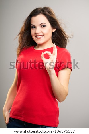 Attractive young woman in a red shirt. Woman shows a sign okay. Hair in motion. On a gray background
