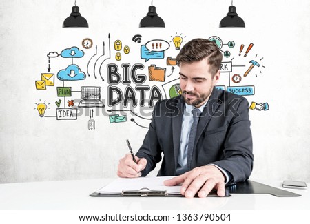 Serious young businessman with beard signing document sitting at table over concrete wall background with colorful big data sketch