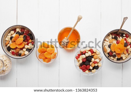 top view of bowls with cereal, dried apricots and berries, honey and nuts served for breakfast on white wooden table