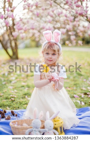 Little cute girl sitting on the grass near the magnolia. A girl dressed as an Easter bunny holds a flower and an egg. Spring.