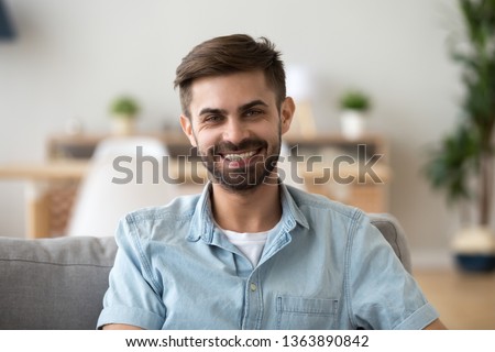 Head shot portrait of happy smiling millennial man sitting on comfortable sofa in living room, looking at camera, successful confident freelancer, excited guy posing for photo at home