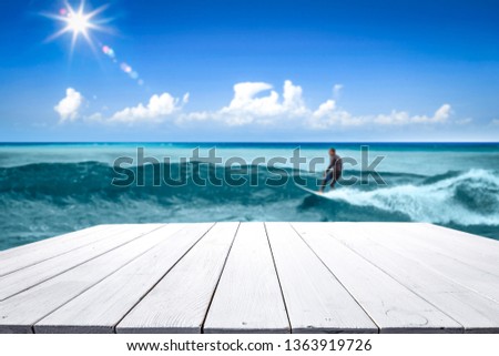 Wooden table background of free space for your decoration. Surfer on board with big waves on ocean. Summer blue sky with sun. 