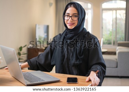 Smiling middle eastern woman wearing abaya and eyeglasses with laptop and smart phone looking at camera while sitting near desk at home