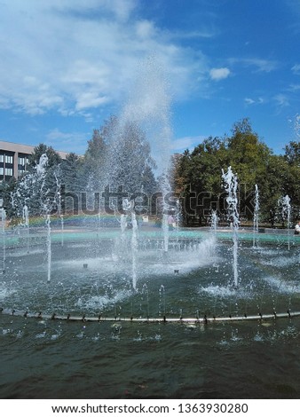 Fountain in the square. Photo of the fountain in the daytime. In the distance, the sky with light clouds, thuja grow and the fountain works.