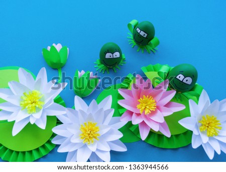 Waterlily flower made of paper. Blue background. Origami hobby. Gentle petal. Marsh with frogs tradition. Egg frog is green. Happy Easter.