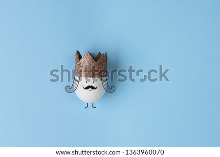 Real egg with gold crown on blue background. White face concept.