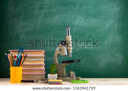 Education and science concept - group of colorful books and microscope on the wooden table in the classroom, blackboard background