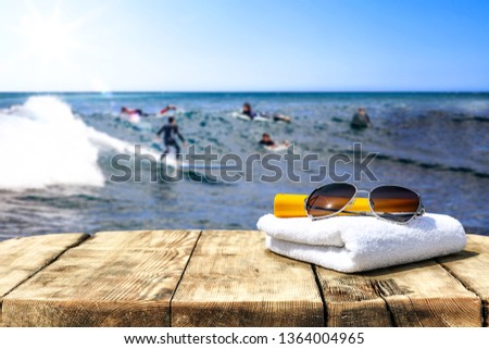 Wooden table background of free space for your decoration. Blurred background of surfers on waves. White towel and sunglasses . Summer time background 