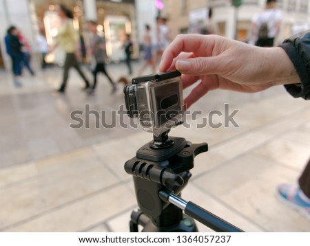 Female hand holding pressing the shutter release button of  an action camera on a tripod in a busy pedestrian street , a few people passing by in the background and a few stores blurry on purpose.