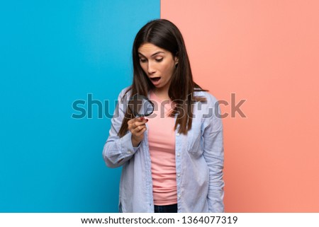 Young woman over pink and blue wall holding a magnifying glass