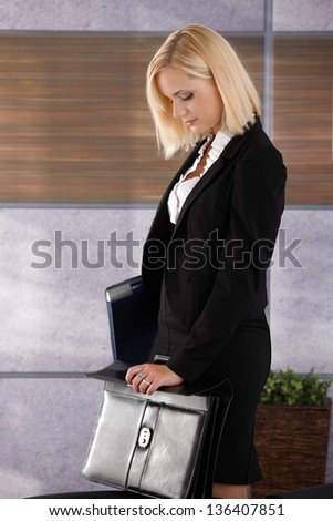Attractive businesswoman standing in office, holding laptop computer and closing business briefcase.