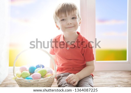 Cheerful smiling boy holding basket full of colorful easter eggs and sitting on the windowsill against the background of a spring green field at sunset or dawn
