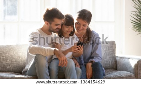Little daughter sitting with parents holding smart phone showing playing games using online apps, mom dad having bought new device teaching kid, people having video call. Modern wireless tech concept