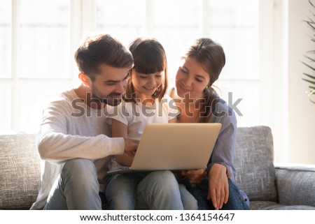 Young couple with little daughter sitting on couch having fun holding computer looking on laptop screen teaching kid use electronic device, surfing web browsing internet shopping buying online concept