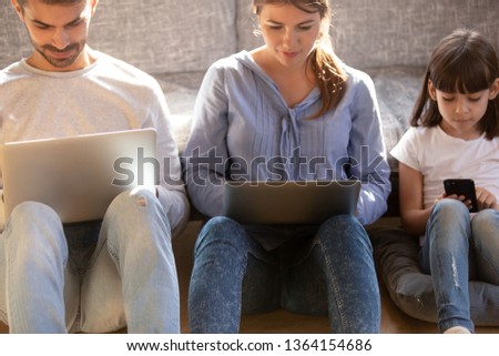 Wife husband and daughter sitting on warm floor leaned on sofa spend time at home family absorbed in electronic devices, working on laptops, play game surfing web, bad habit, gadgets addiction concept