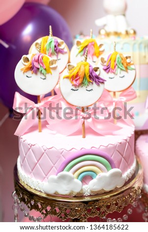 Pink cake with marzipan, with a rainbow and white clouds, and cookies on sticks with the image of a unicorn.